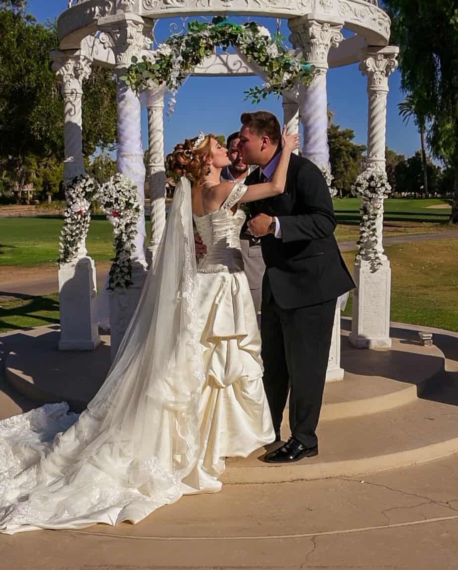 A bride and groom share a romantic kiss during their wedding ceremony outdoors, surrounded by beautiful floral decorations and a scenic park setting.