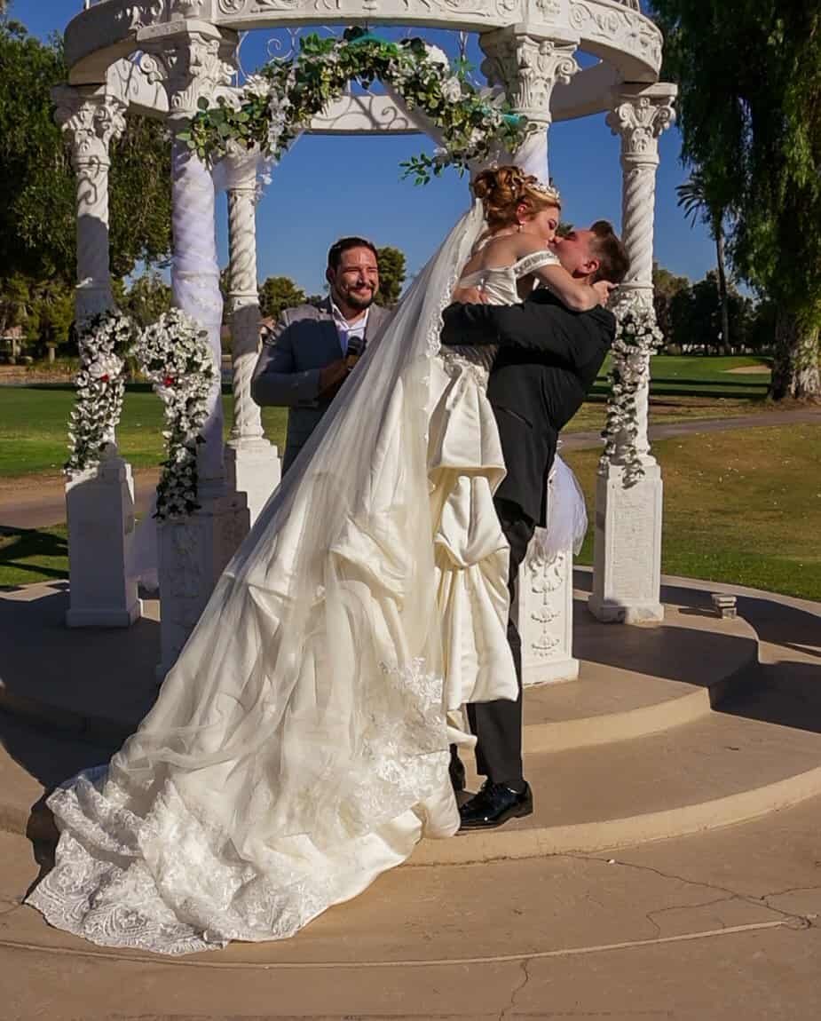 A joyful bride and groom share a romantic moment during their outdoor wedding ceremony beneath a floral-adorned gazebo.