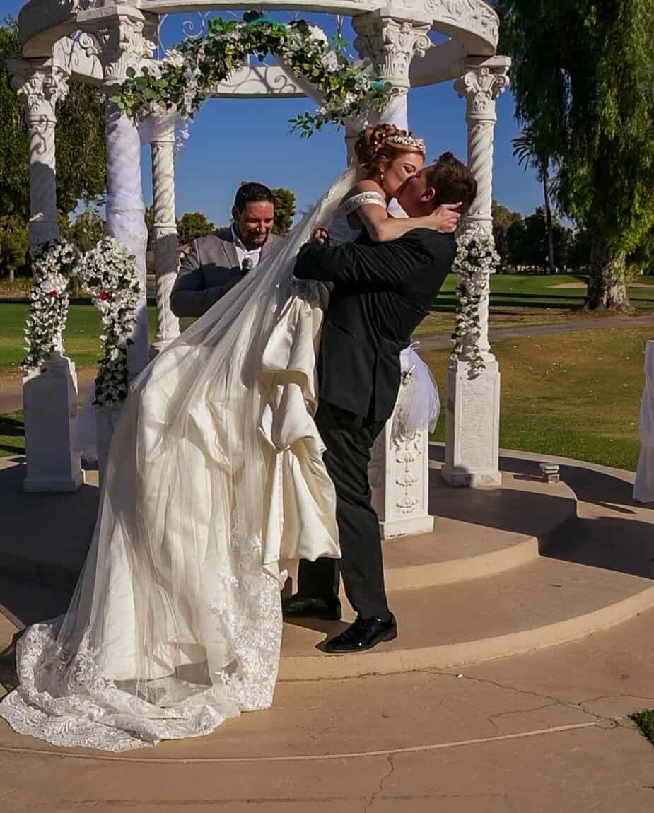 A romantic outdoor wedding scene featuring the bride and groom embracing during their ceremony at a beautifully decorated gazebo.