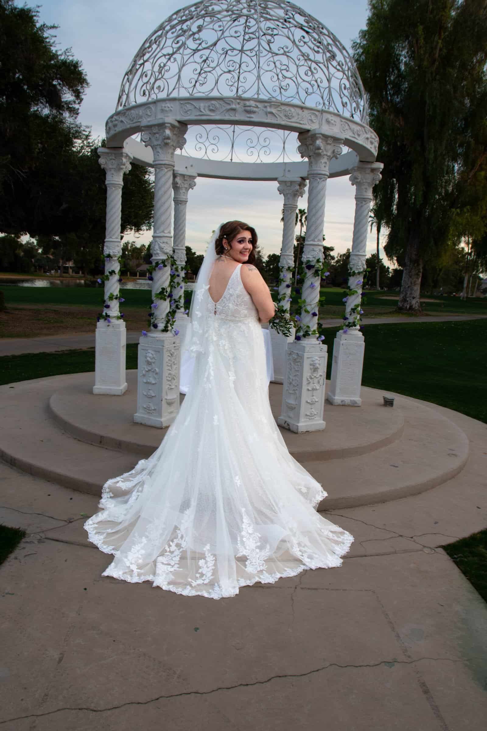 Beautiful bride in wedding dress posing at decorative gazebo in park setting.