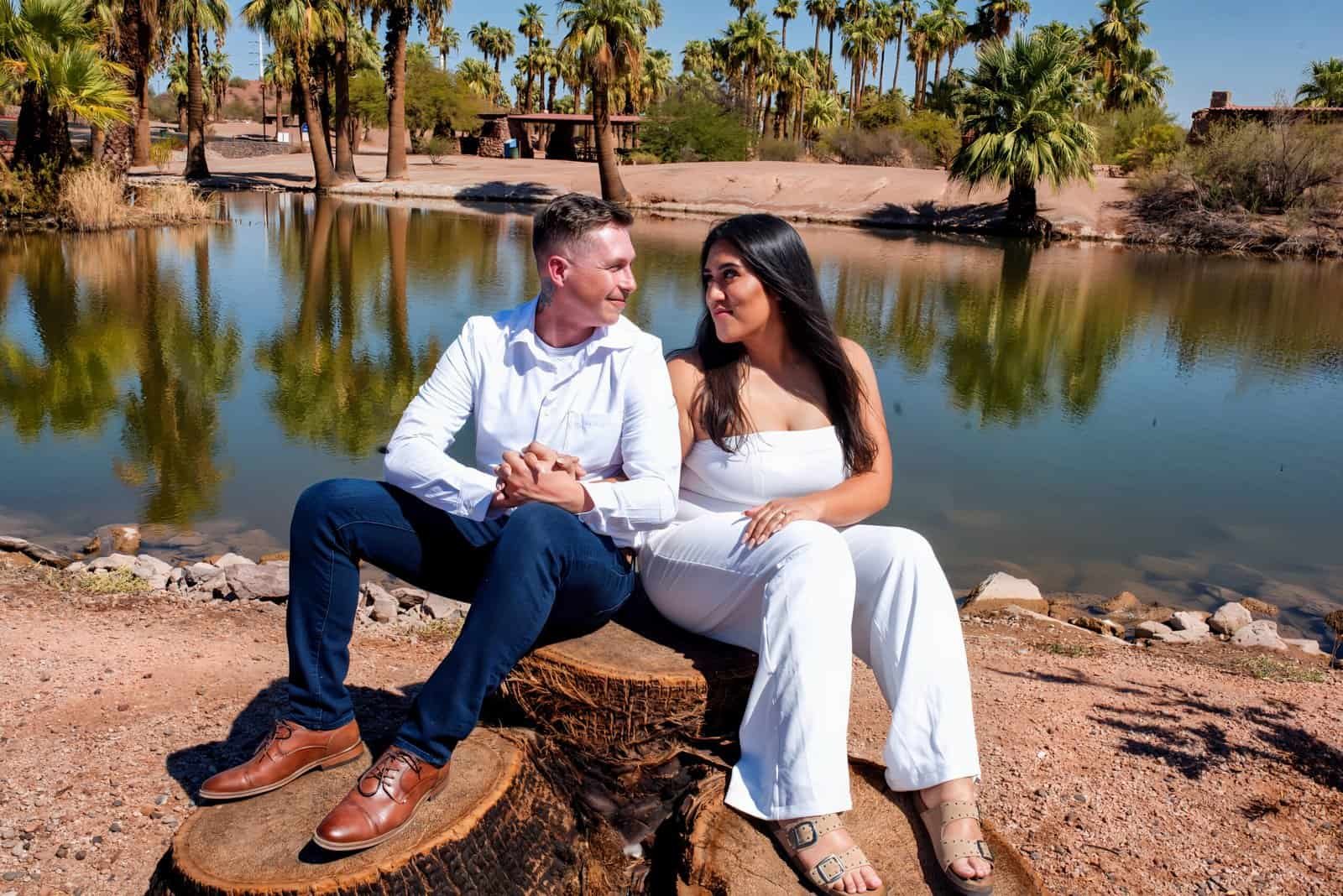 A happy couple enjoying a romantic moment by the water in a lush, scenic outdoor location with palm trees and calm waters.