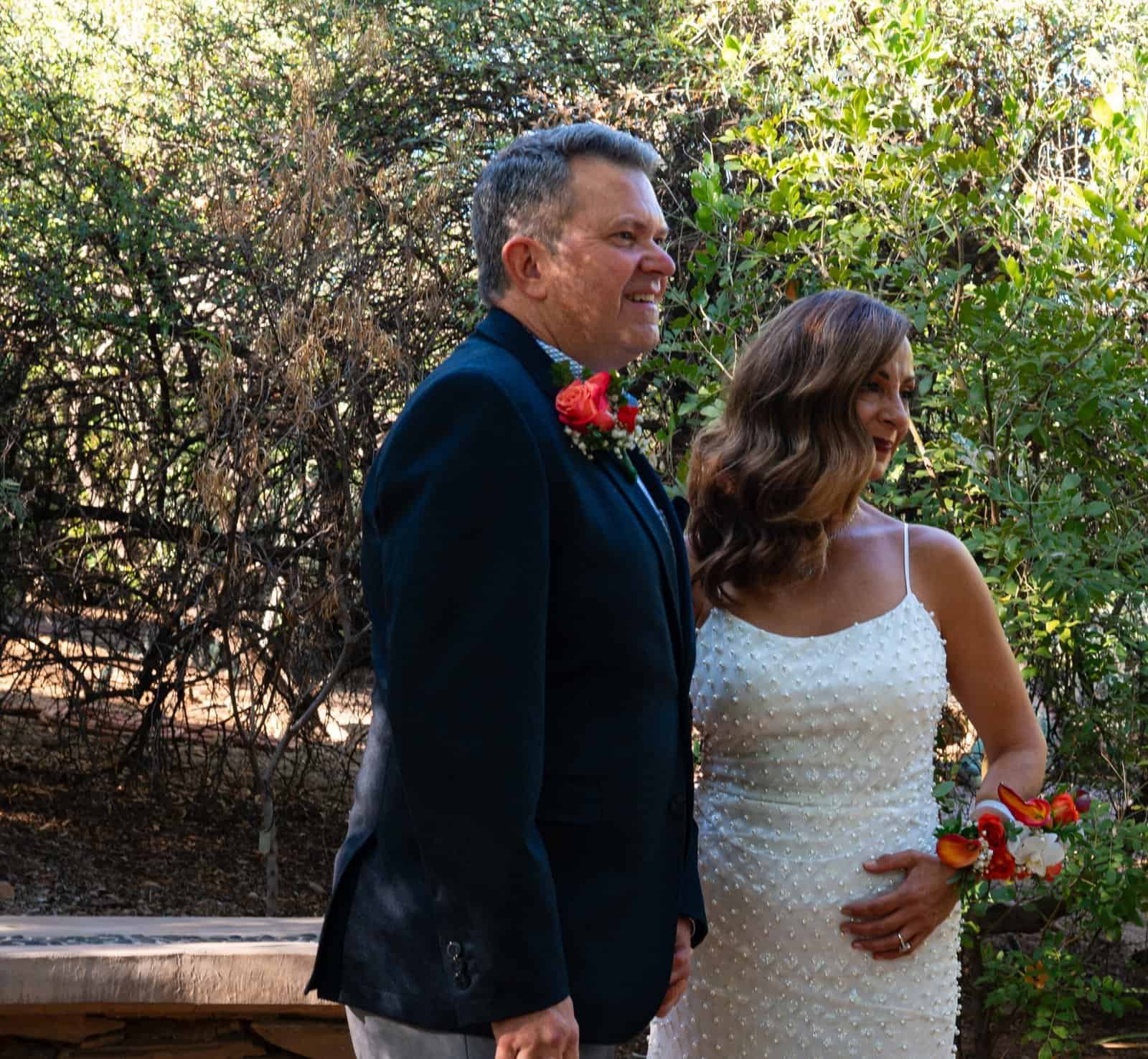 A bride and groom stand together outdoors, celebrating their wedding surrounded by greenery.