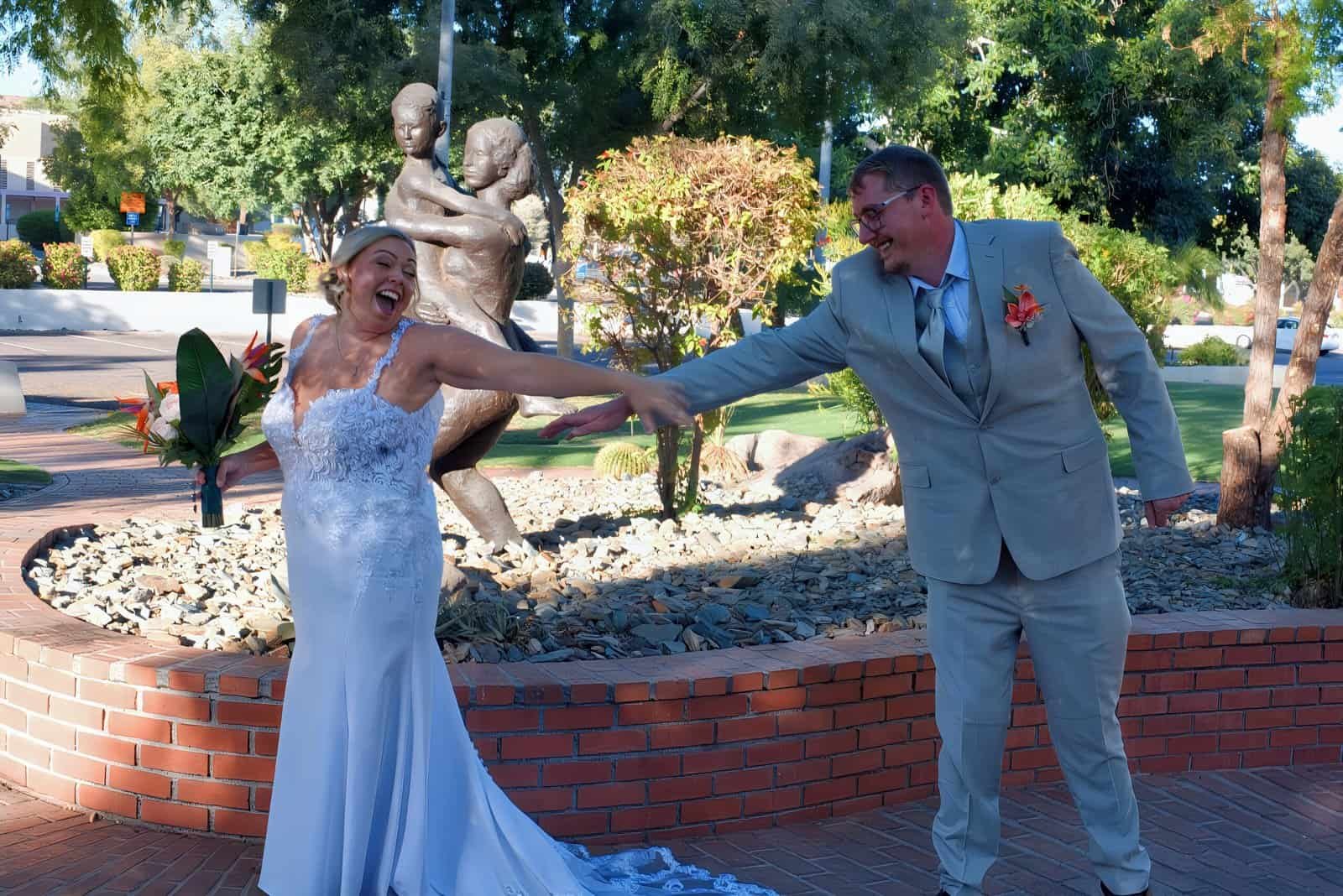 A bride and groom enjoying a lively dance in a scenic park with trees and a statue in the background.