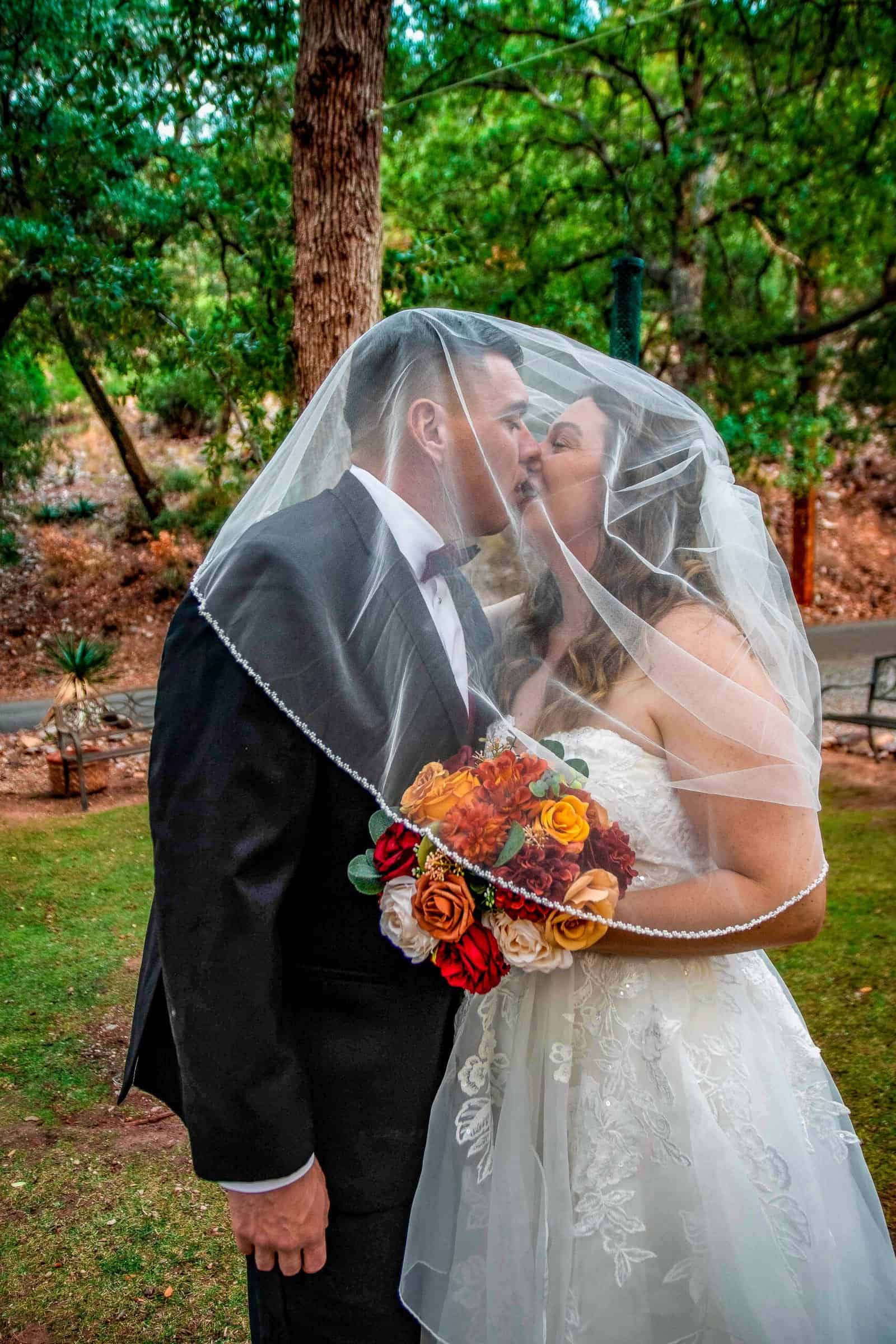A bride and groom kiss tenderly under a veil in a lush outdoor setting, capturing a heartfelt wedding moment.