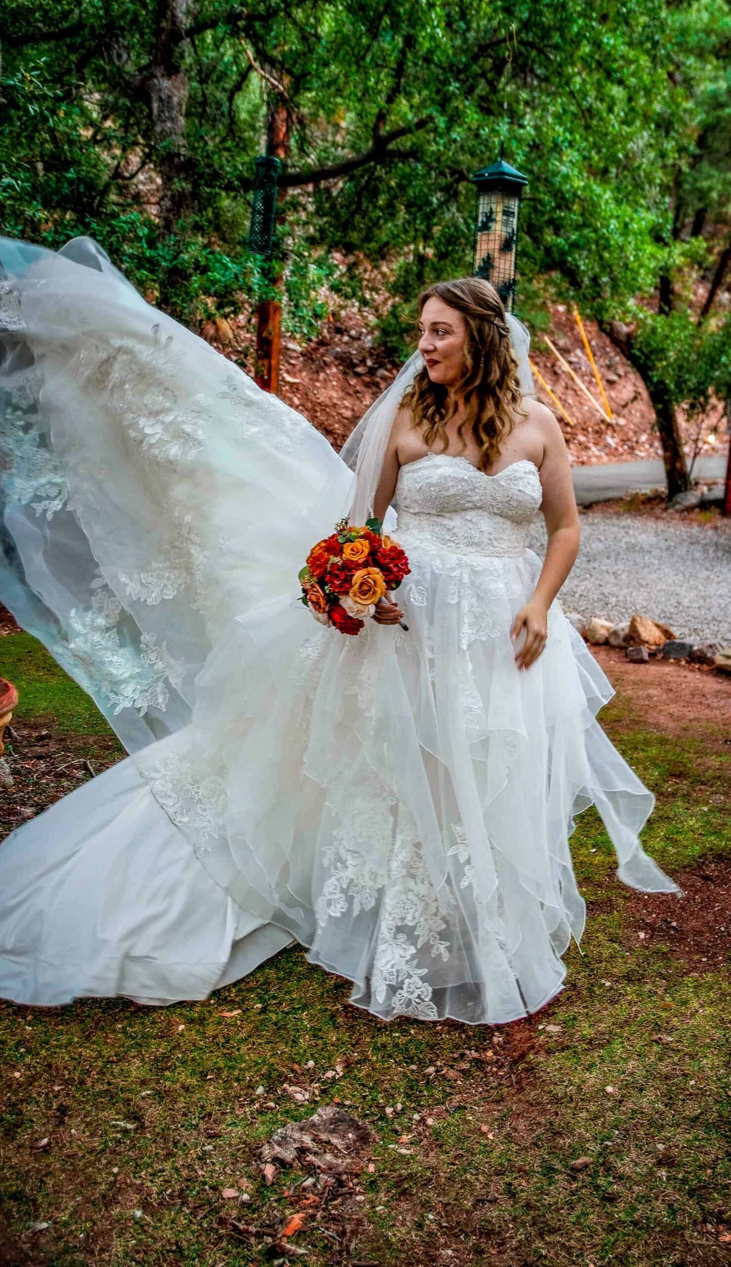 A bride in a beautiful wedding dress holding a vibrant bouquet, standing in a scenic outdoor location surrounded by trees.
