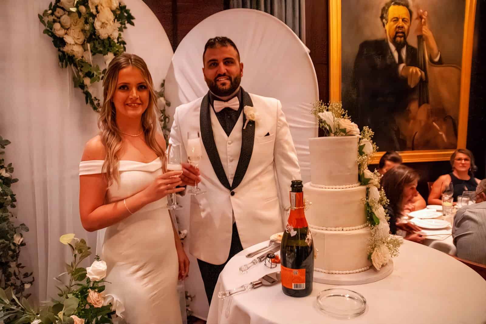 A bride and groom stand together at their wedding reception, smiling and holding glasses of champagne near a decorated cake.