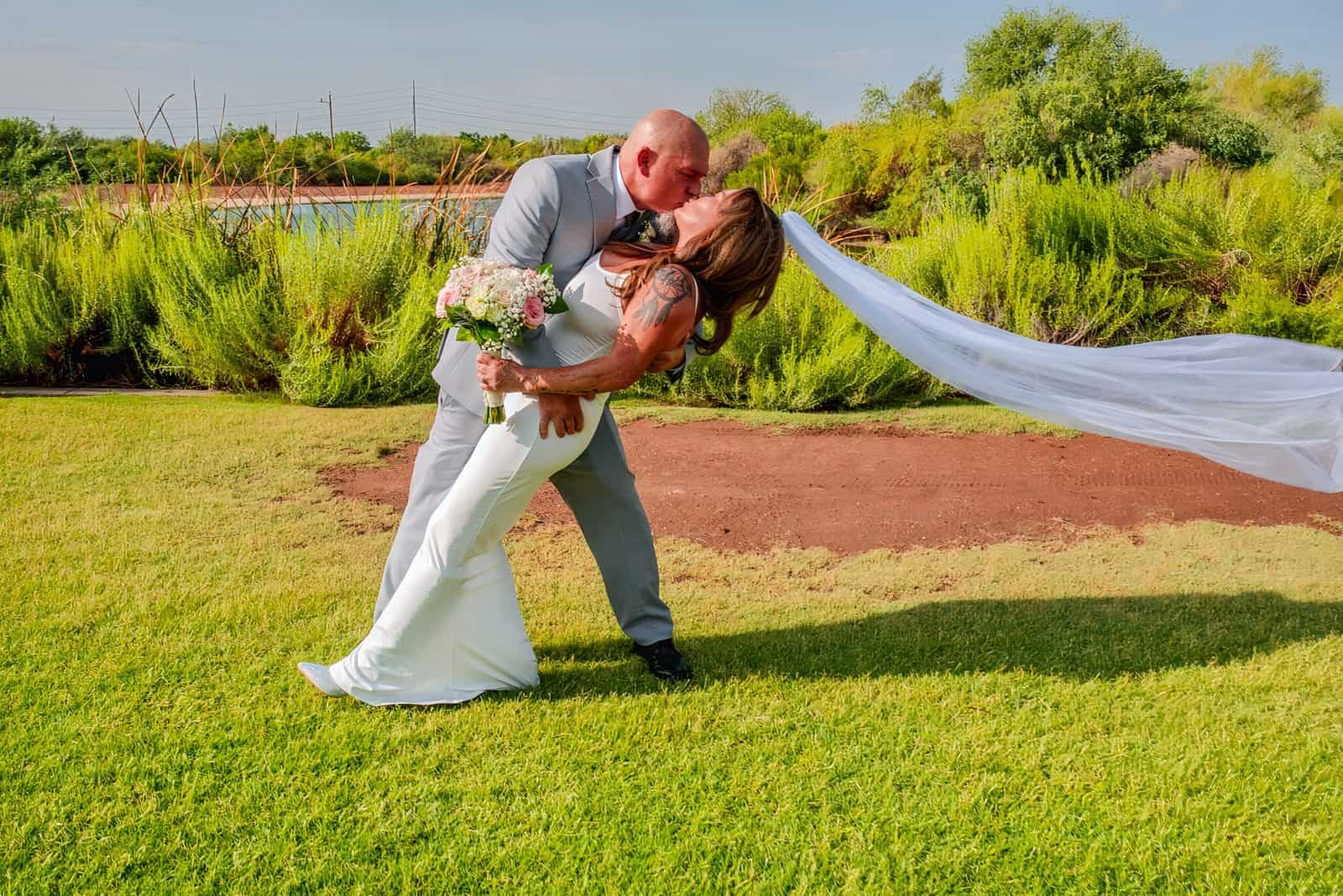 A couple in wedding attire sharing a kiss during their outdoor wedding celebration on a sunny day.