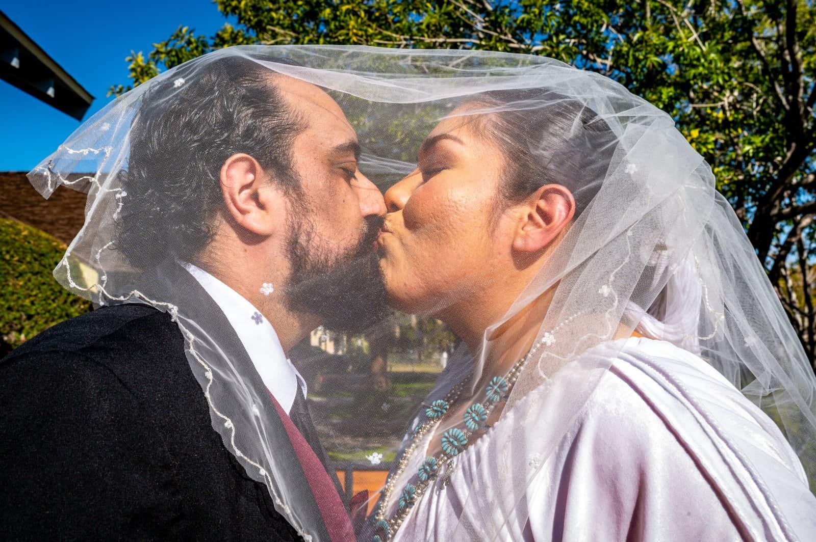 Romantic moment of a bride and groom sharing a kiss under a veil in a scenic outdoor setting.