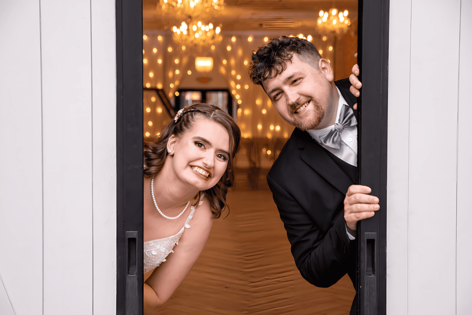 Joyful bride and groom peeking through a door at their wedding reception, capturing a candid moment of happiness.