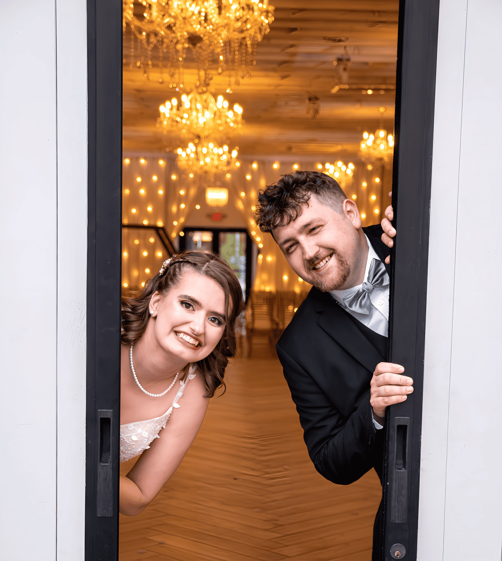 Joyful bride and groom smiling and looking through a door at their wedding reception with warm, festive lighting.