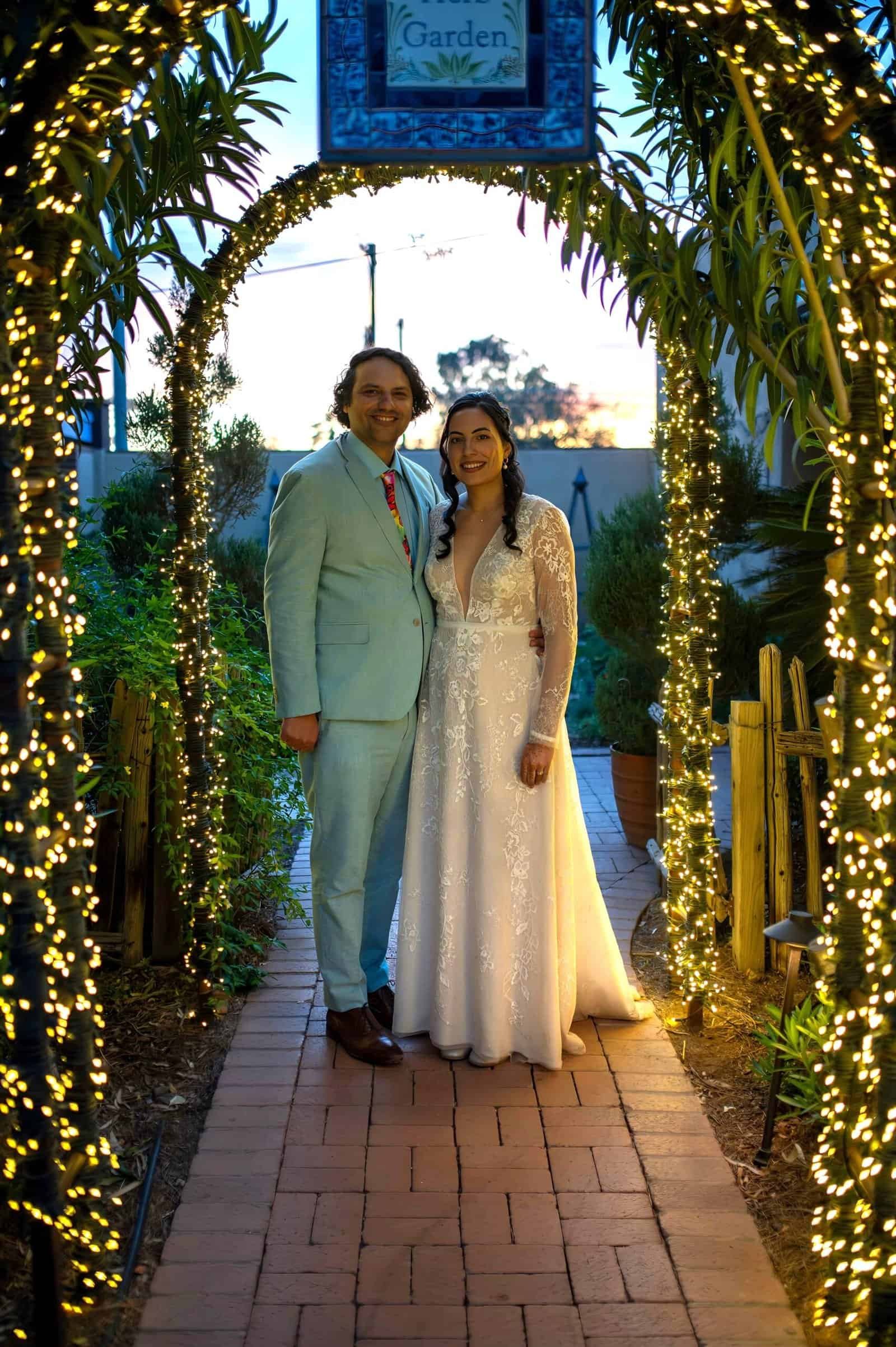 A joyful bride and groom pose together beneath a beautifully lit floral arch at sunset, capturing a memorable moment of their wedding celebration.
