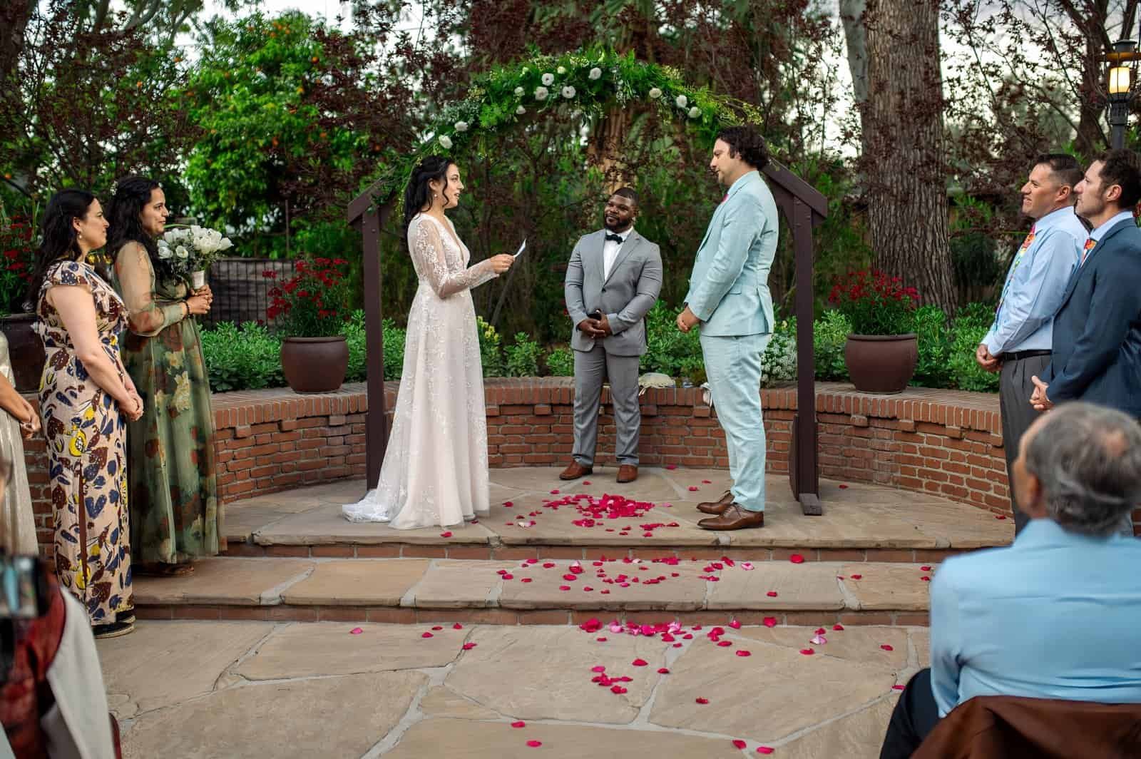 Beautiful outdoor wedding with guests witnessing vows, rose petals on the ground, and lush greenery in the background.