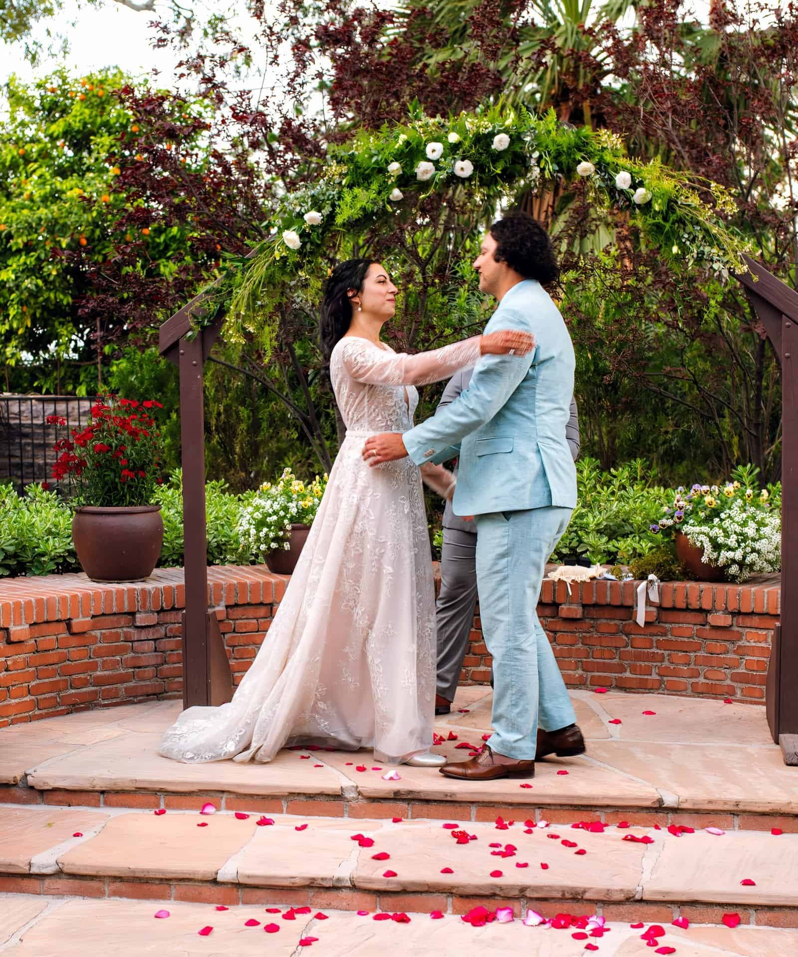 Beautiful outdoor wedding ceremony with bride and groom sharing a special moment under a floral arch, surrounded by lush greenery.