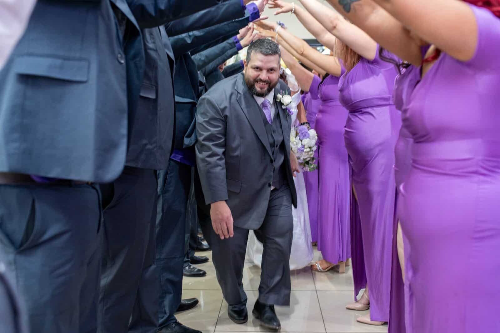 Happy groom surrounded by bridesmaids in purple dresses forming a celebratory tunnel.