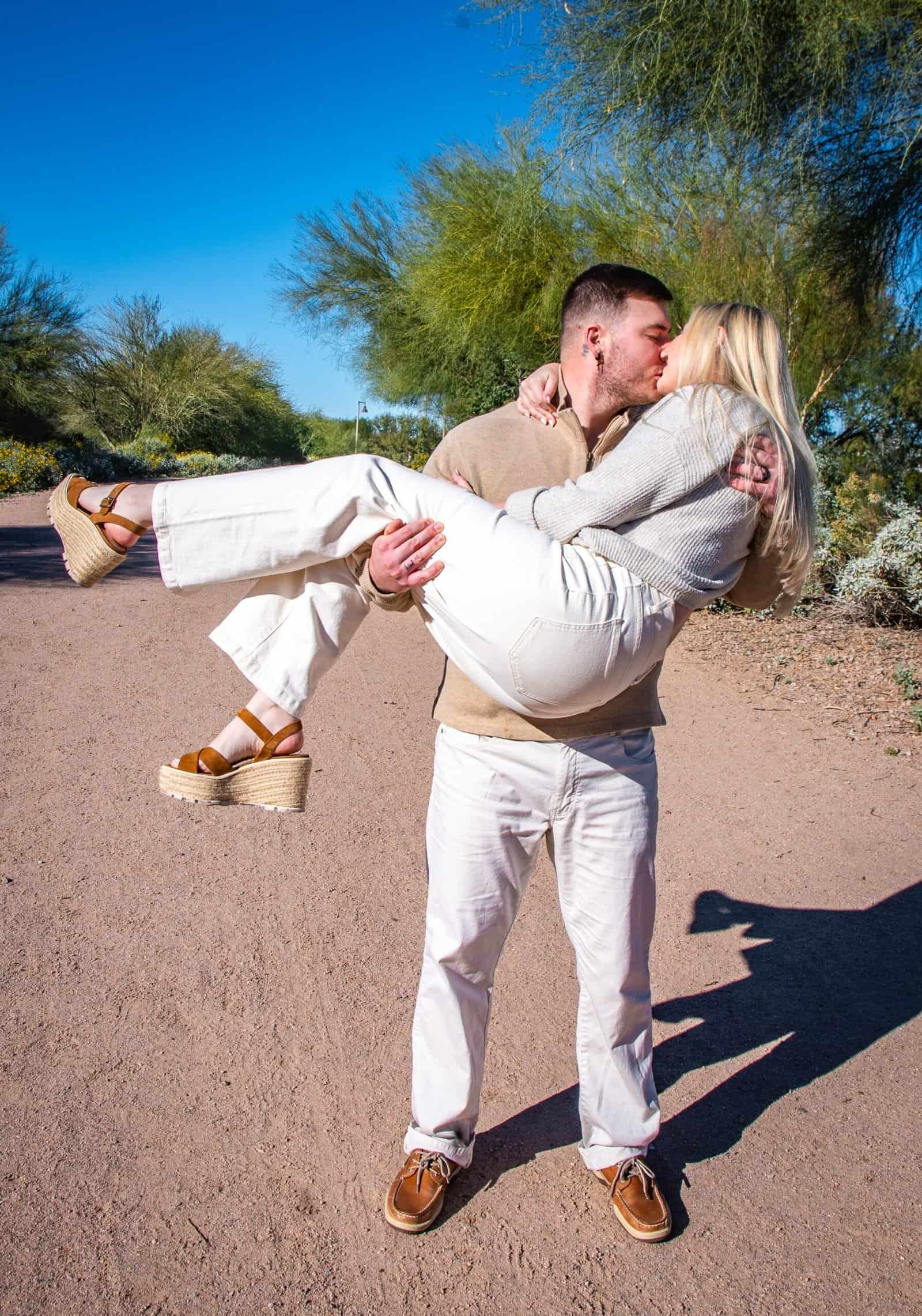 A man lifts a woman in his arms, both dressed in light-colored clothing, surrounded by desert plants and clear blue sky.