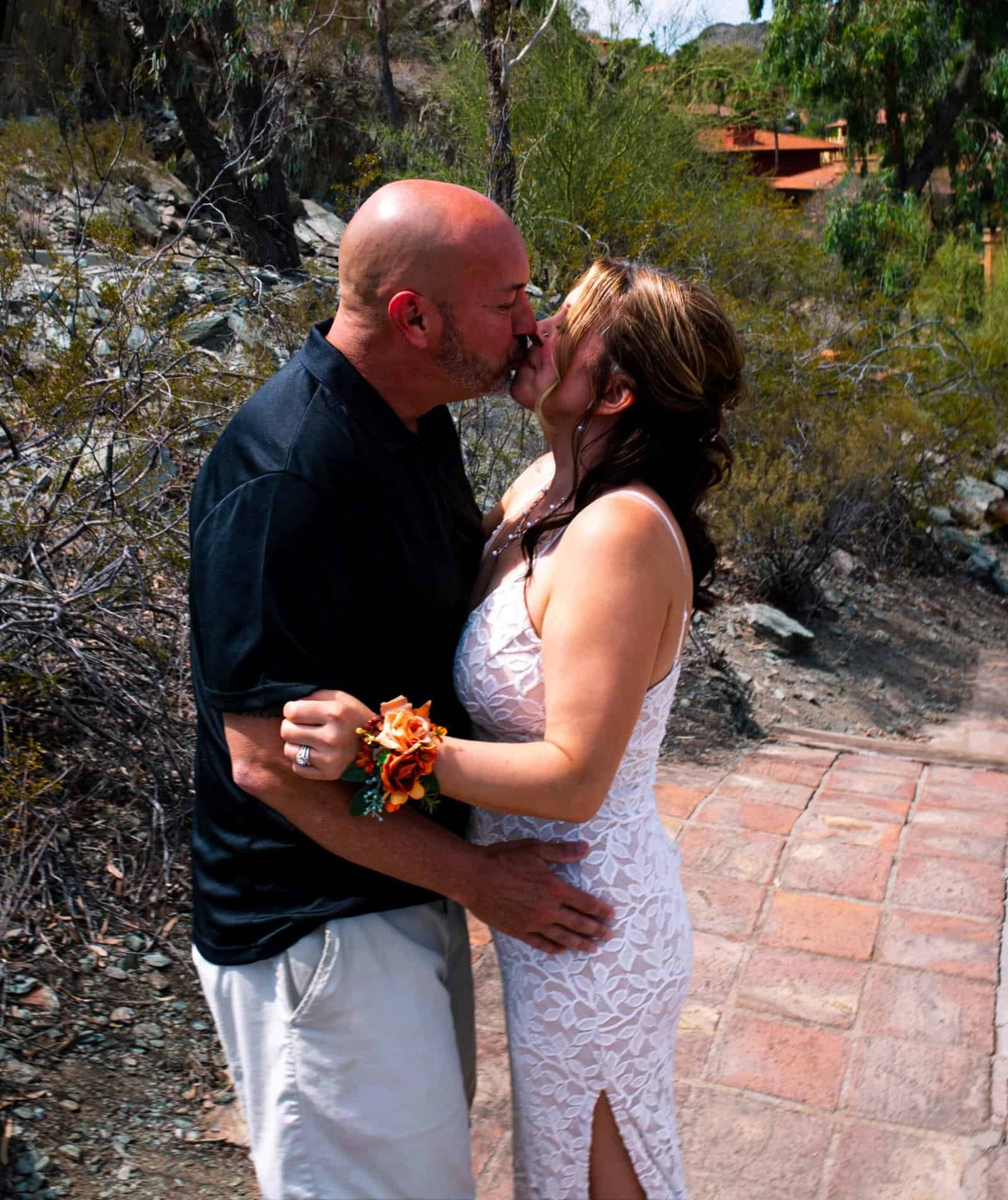 A romantic couple shares a tender kiss during their outdoor wedding, surrounded by nature and warm sunset lighting.