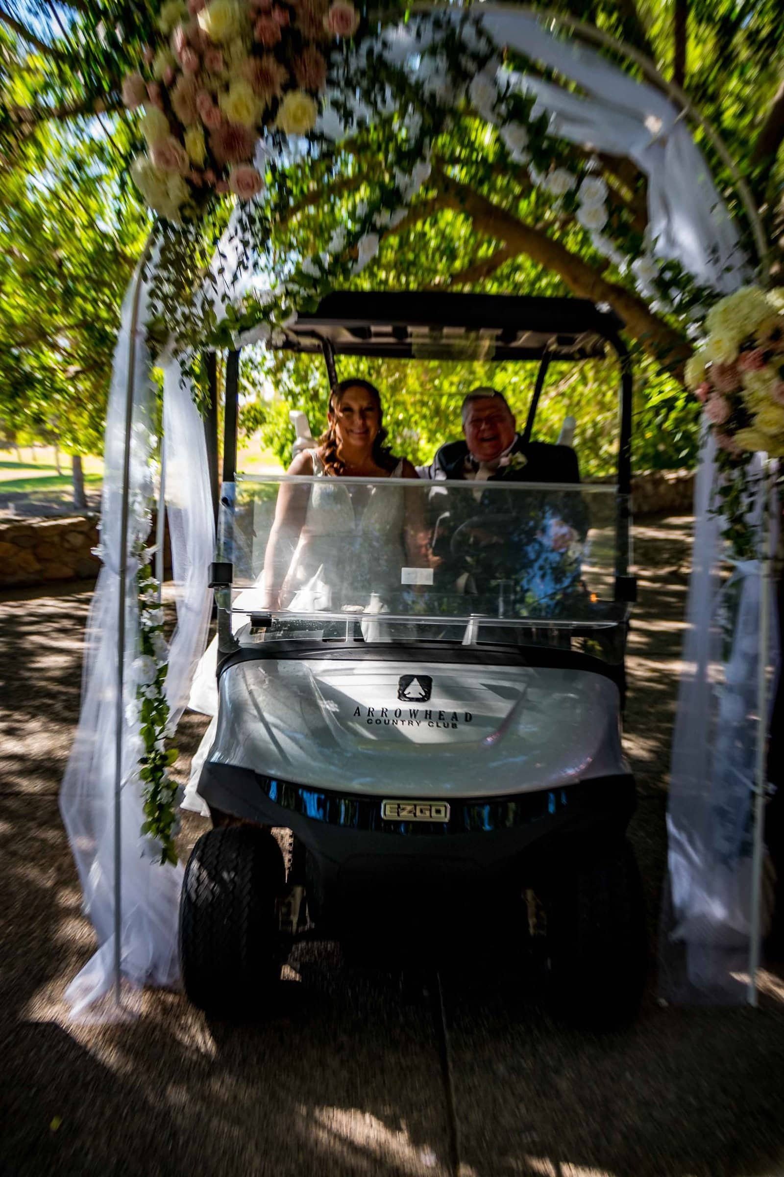 Bride and groom riding in a decorated golf cart under a floral archway during their wedding celebration.