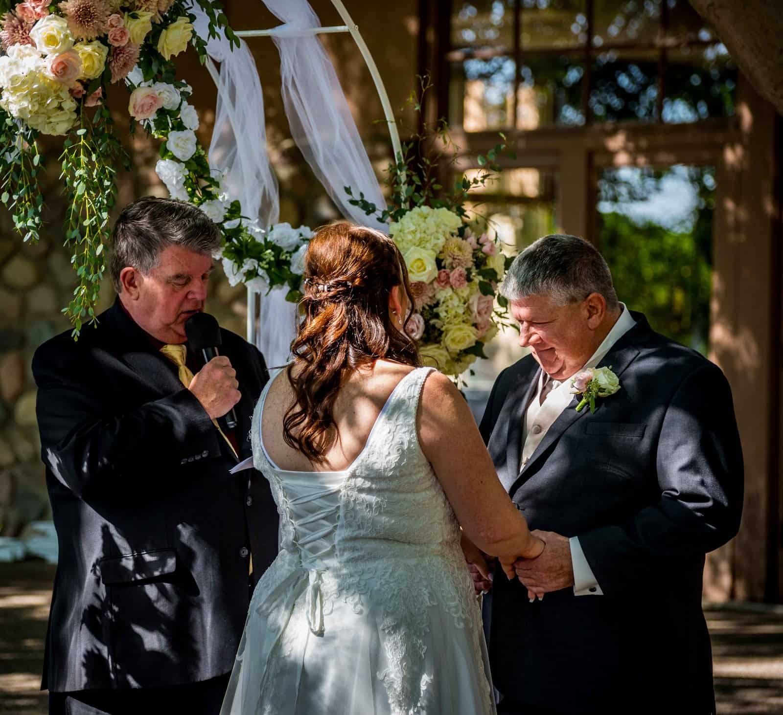 A touching wedding moment with the bride and groom holding hands during their vows, surrounded by floral decorations.