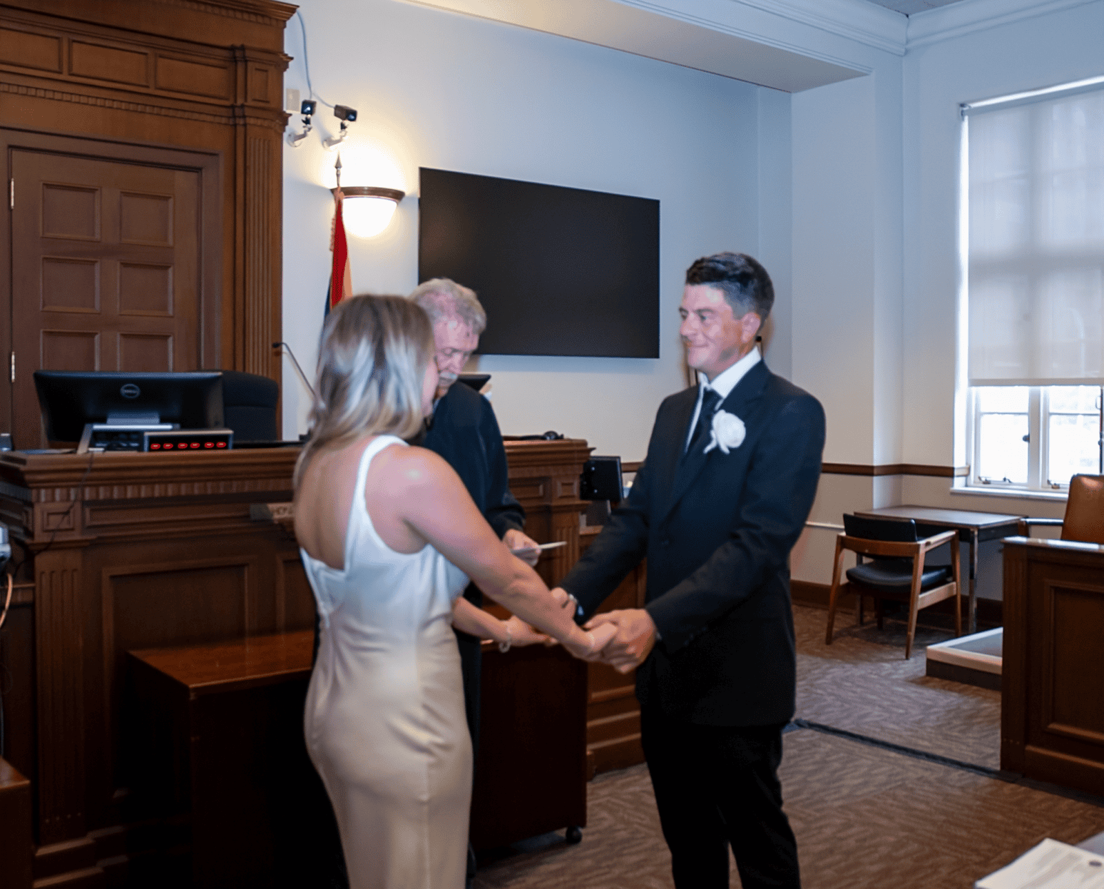 A bride and groom hold hands and exchange vows during their wedding in a courtroom setting.