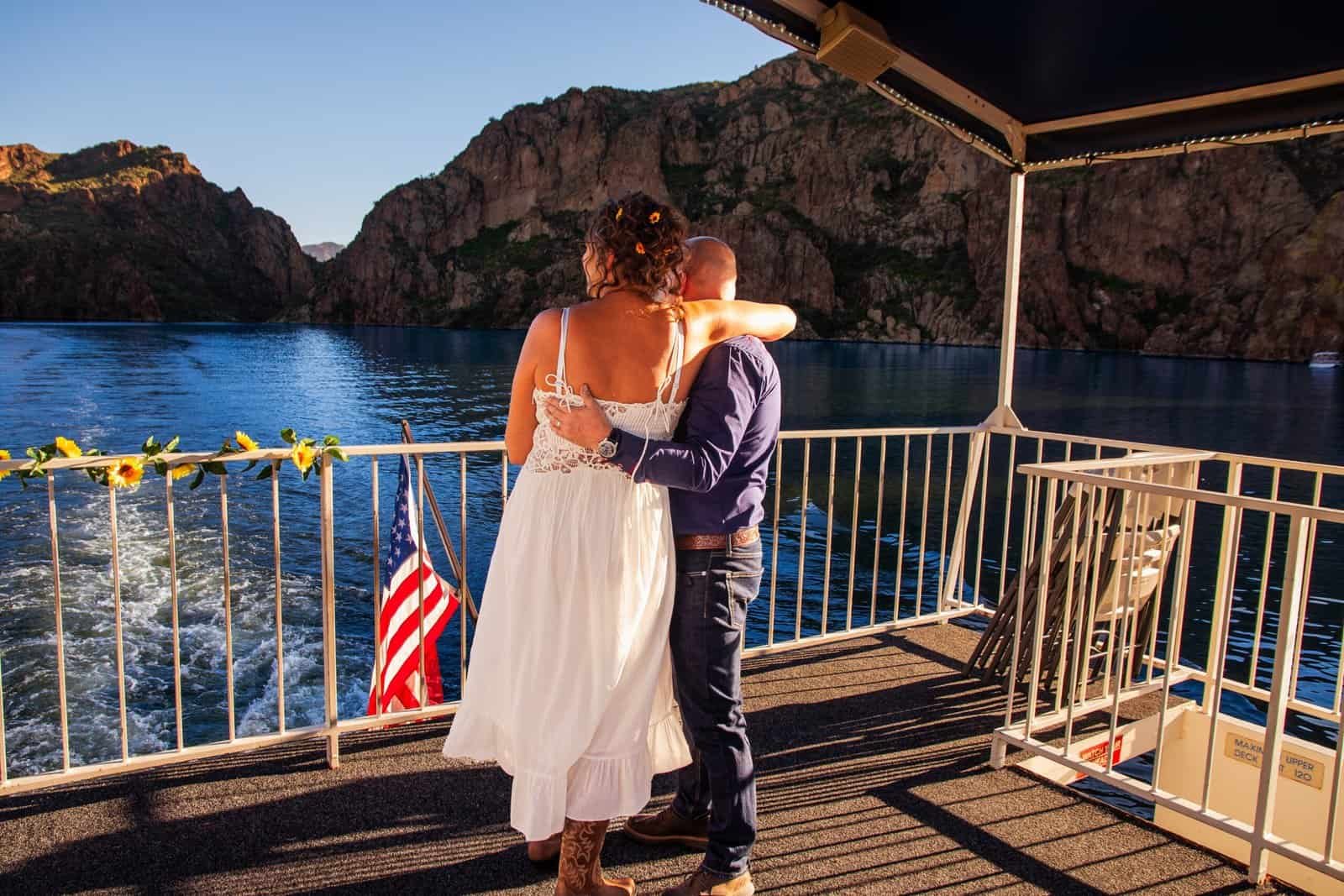A couple shares a tender moment on a boat deck overlooking a beautiful lake surrounded by mountains during sunset, capturing love and adventure.