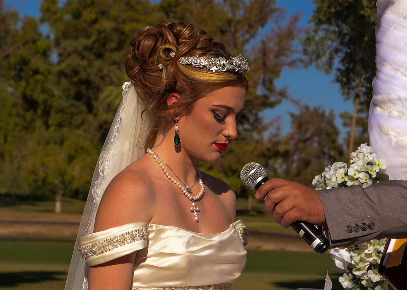 A bride in a satin wedding dress and pearl jewelry stands with eyes closed as she recites vows during her outdoor wedding ceremony.