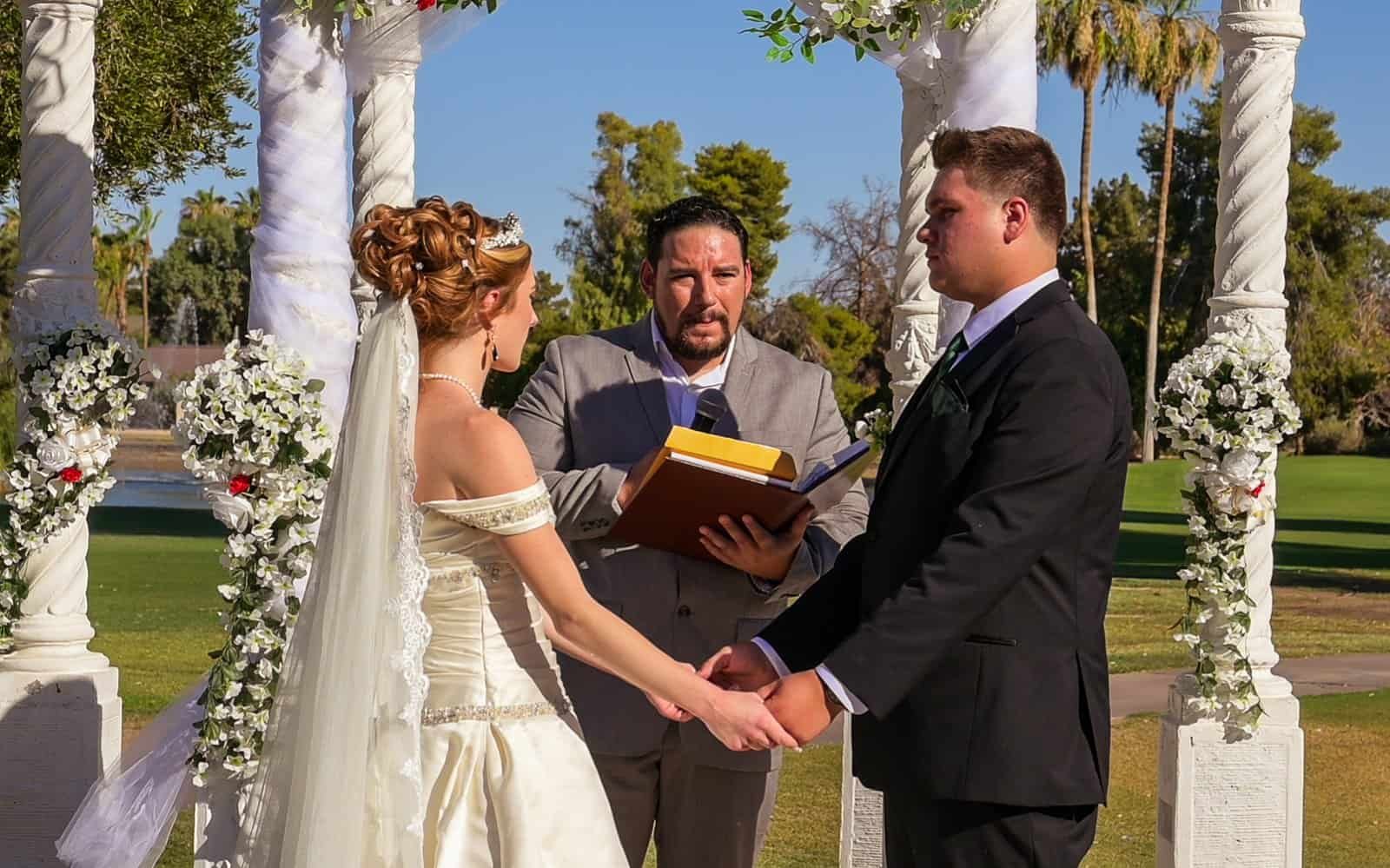 A bride and groom holding hands during their outdoor wedding ceremony, officiated by a person reading vows.