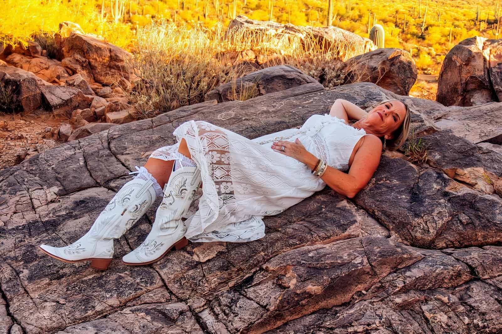 Relaxed woman in a white dress and cowboy boots lying on rocks during sunset in a scenic outdoor location.