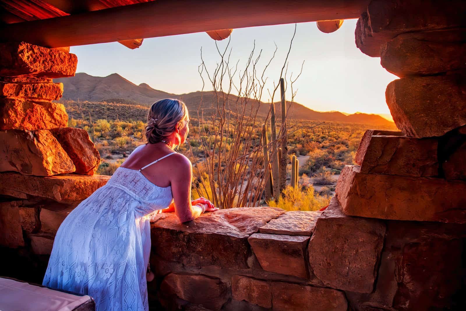 A woman in a white dress enjoying a stunning desert sunset view from a rustic stone window, with mountains and cacti in the background.