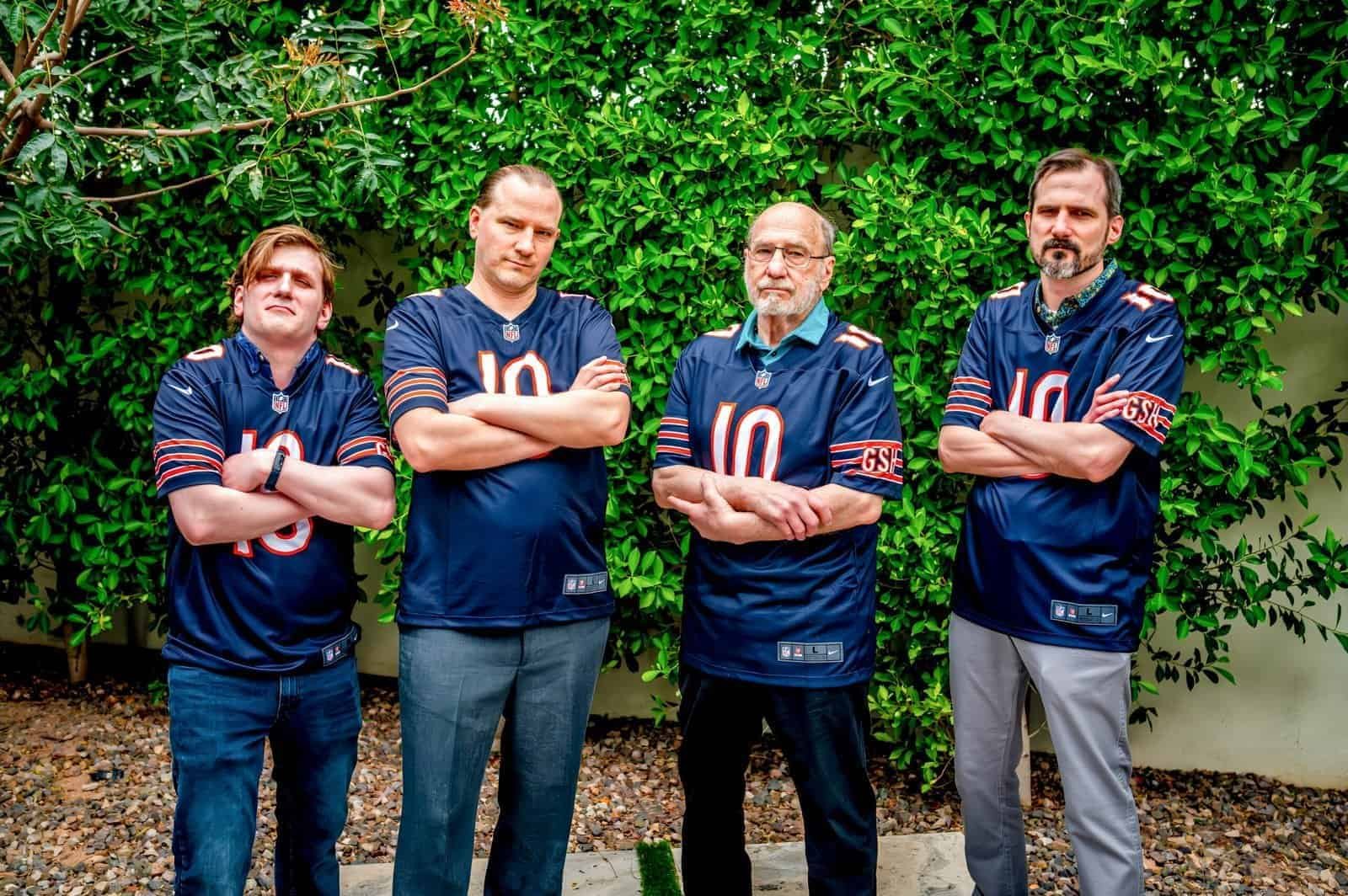 Group of four men in sports jerseys posing confidently outdoors with lush greenery in the background.