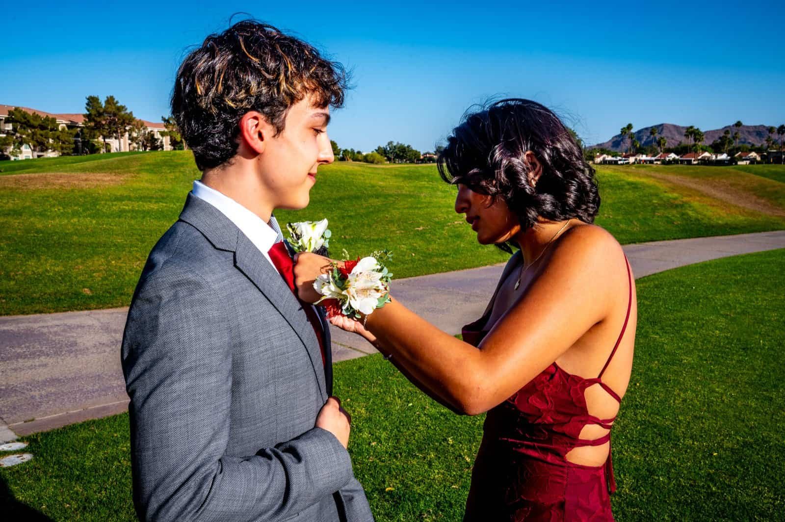 A young man in a suit and a woman in a dress share a heartfelt moment during an outdoor event in a lush park.
