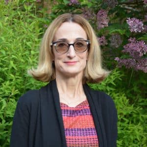 Smiling woman with blonde hair standing in front of bushes an flowers