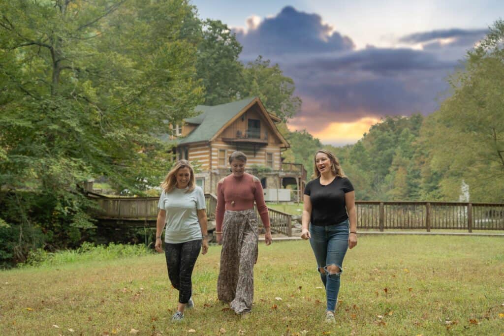 Three women walking in a lush green park near a rustic cabin at sunset.