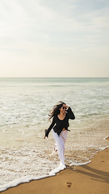 Kvinde i sorte solbriller og hvidt tøj ved stranden, nyder havudsigten.
