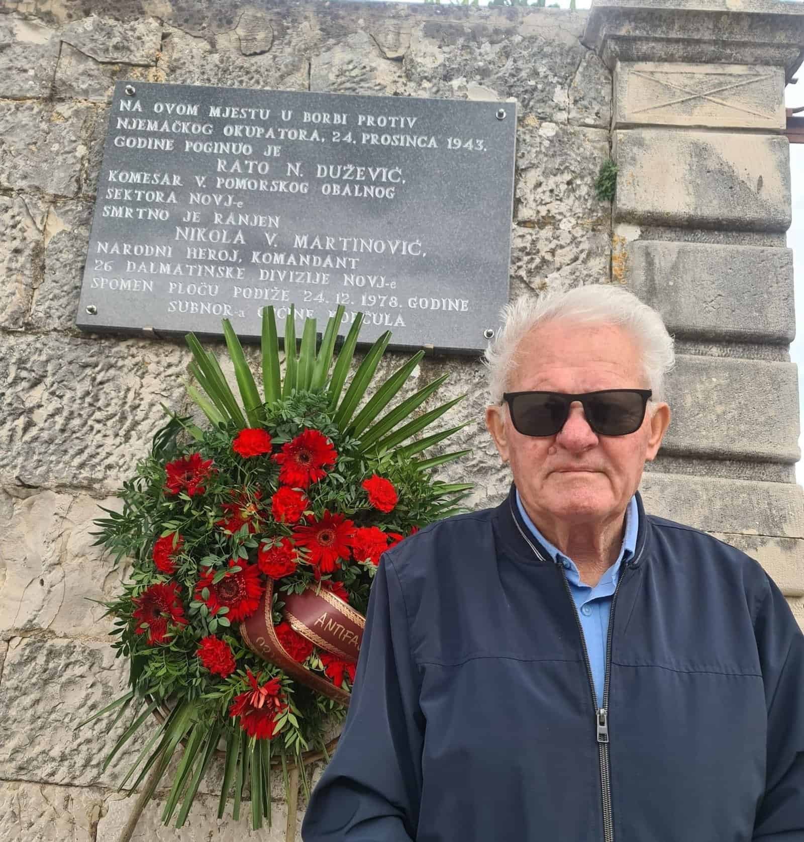 Muško sa sivom kosom, sunčanim naočalama i plavom košuljom ispred spomen-ploče i vijenaca na memorijalnom mjestu.
