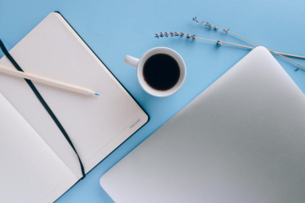 Suite Web Design Blog WordPress Maintenance and Support Services Image of open notebook with blue pencil with a cup of coffee and a closed laptop beside it with a blue background and two sprigs of lavendar layed out flat.