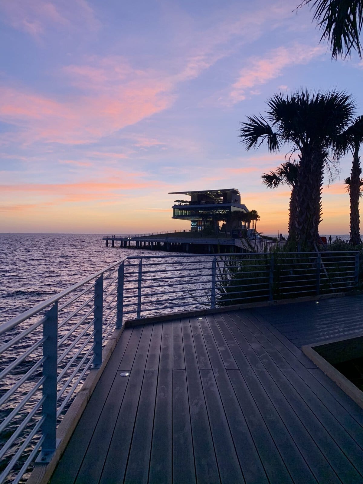 Pastel sunset glowing over St. Pete Pier, with palm tree silhouettes and calm water.