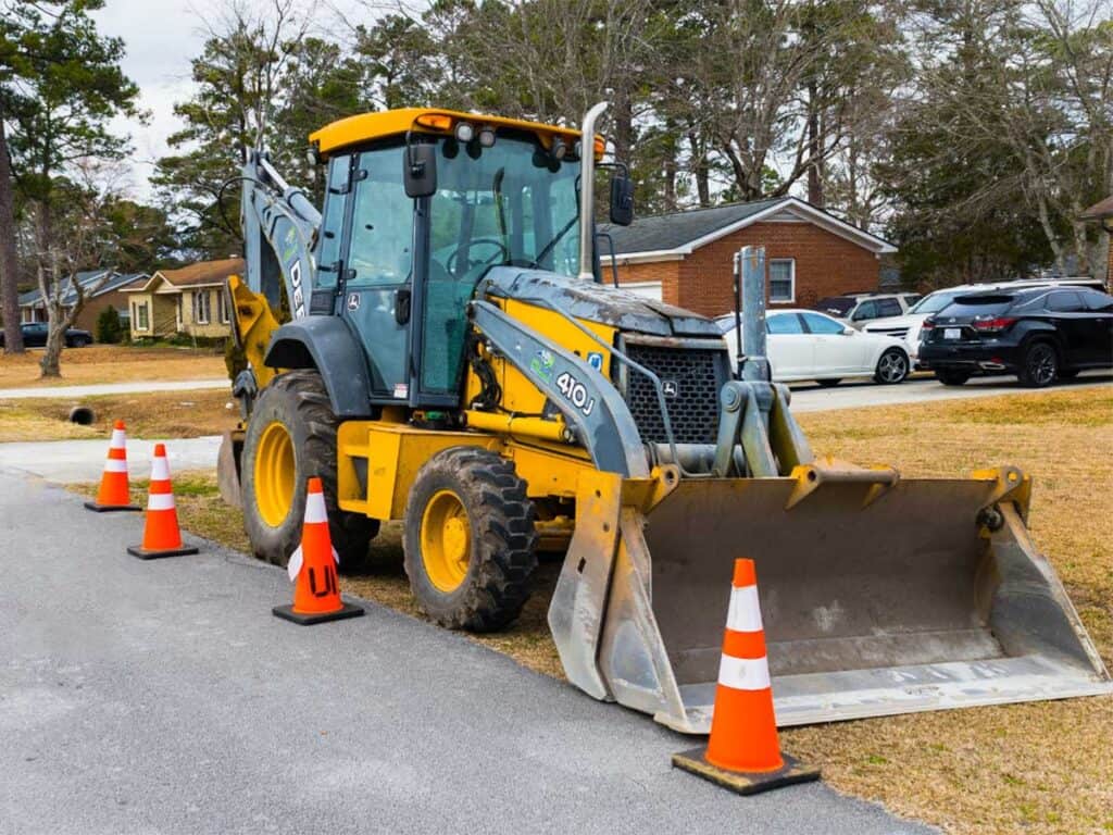 A yellow backhoe loader from Superior Excavating is parked on the side of a residential street, surrounded by orange traffic cones. Houses, trees, and parked cars are visible in the background.