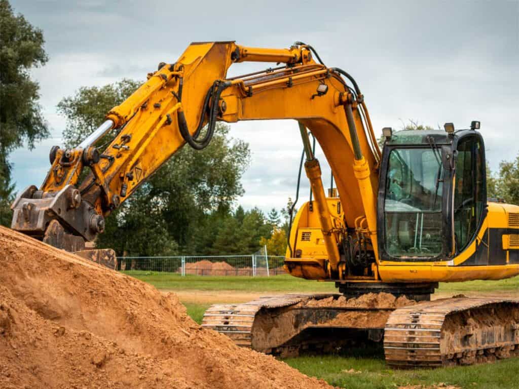 A large yellow excavator from Superior Excavating Auburn Hills MI sits on grass beside a mound of dirt, with trees and a cloudy sky in the background.