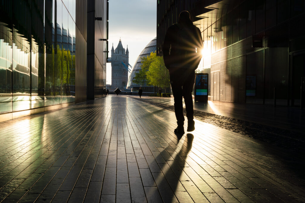 A silhouette of a person walking along a modern city walkway at sunset, with sun rays bursting between buildings and Tower Bridge visible in the background.