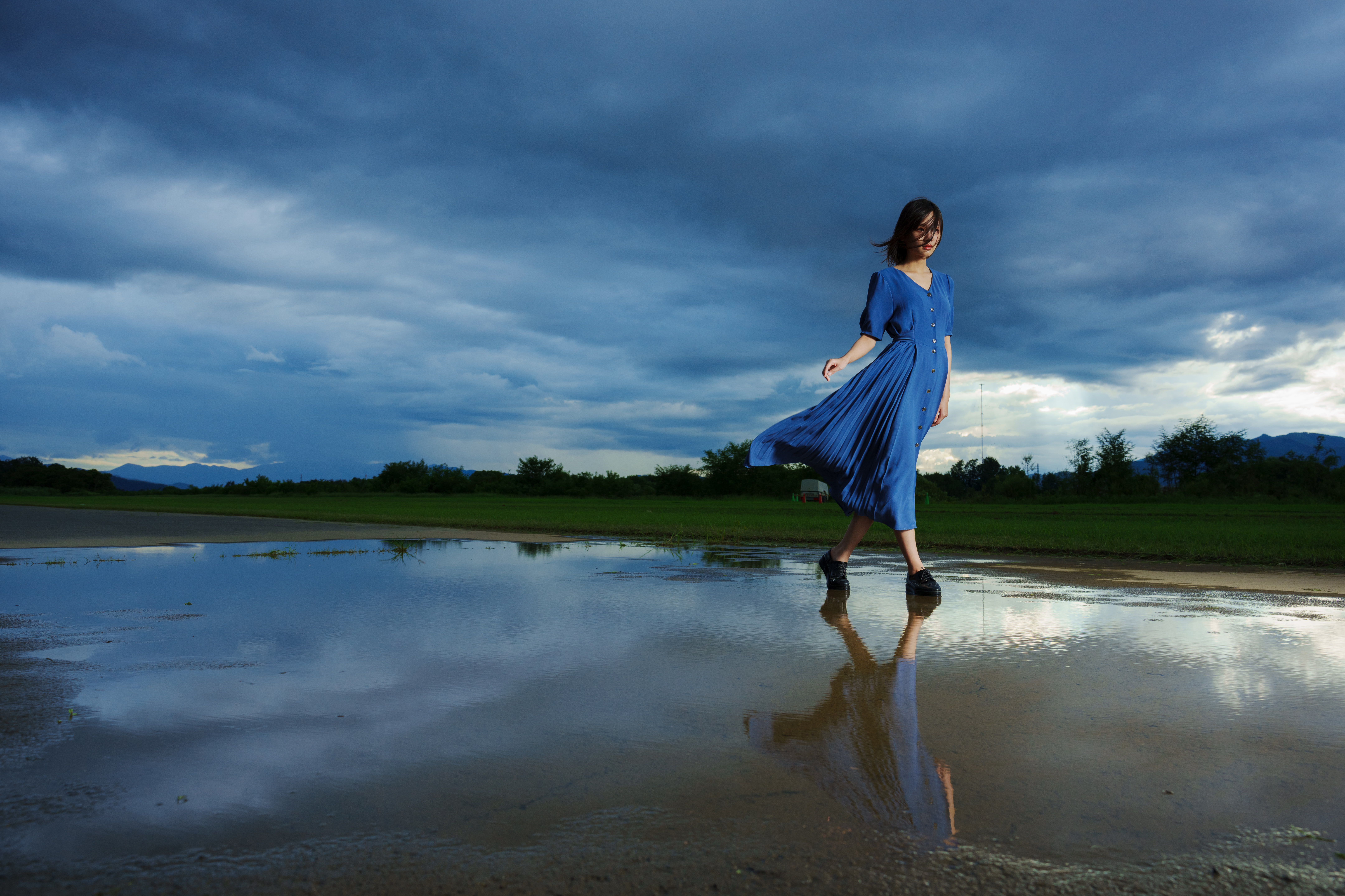 A woman in a blue dress walking across a reflective puddle under dramatic skies, captured with sharp detail and vibrant colors using Tamron lenses for Sony A7R series cameras.