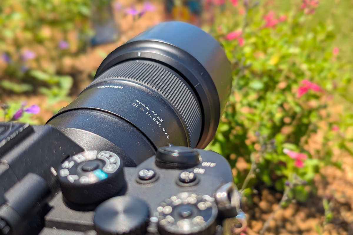 Close-up of a Tamron 90mm F/2.8 Di III Macro VXD lens mounted on a Sony mirrorless camera, set up in a flower garden for photographing insects and macro photography.