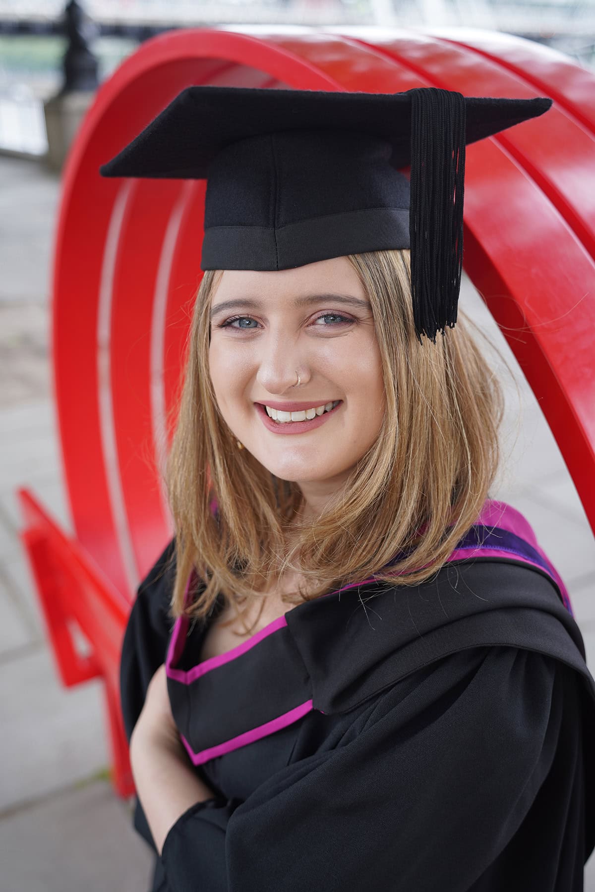 Un diplômé souriant, en bonnet et robe, posant devant une sculpture rouge, capturé avec l'un des meilleurs objectifs de photographie événementielle pour les remises de diplômes.