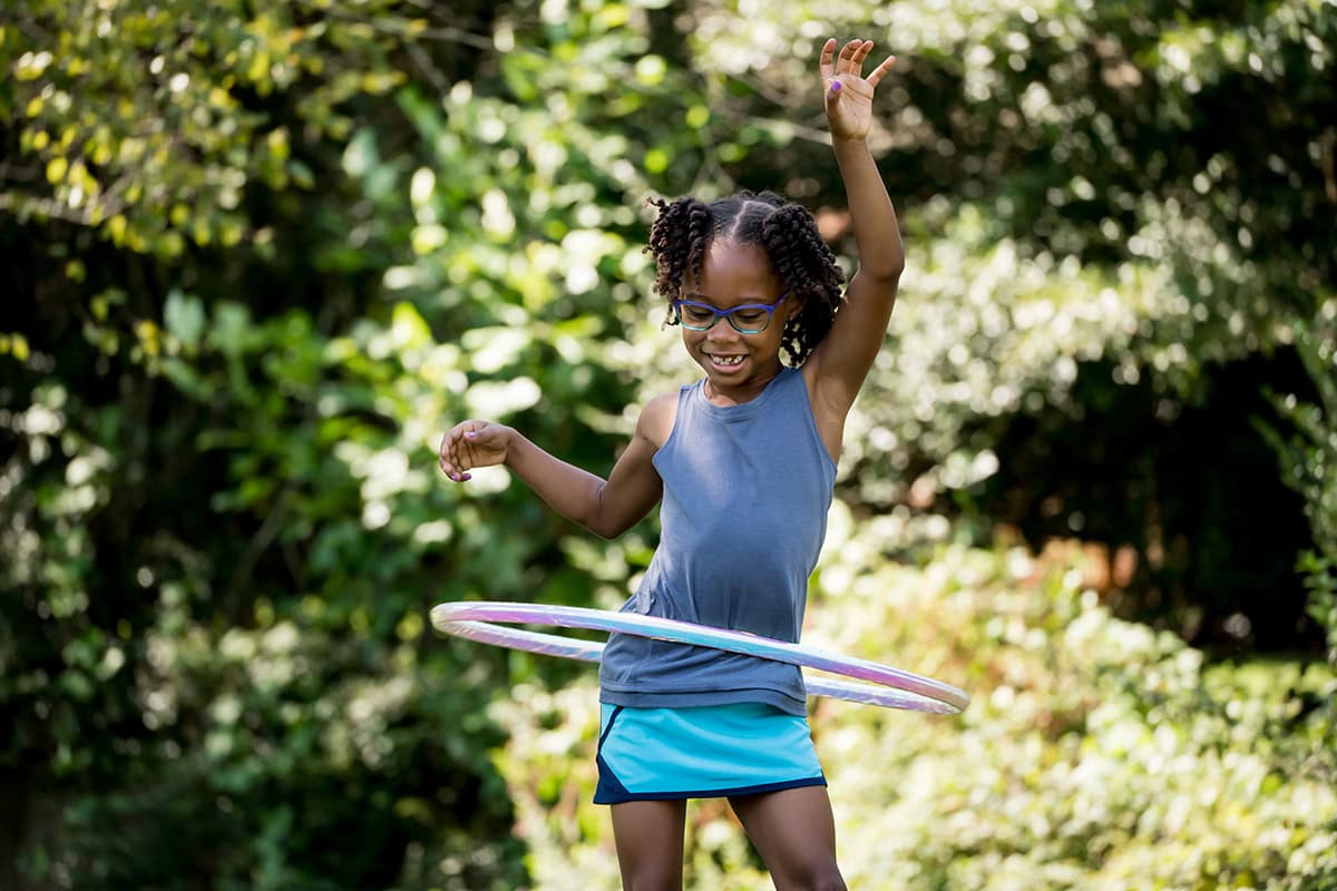 Une jeune fille à lunettes fait joyeusement tourner un hula hoop autour de sa taille en plein air, mettant en évidence l'amusement et l'énergie qui rendent la photographie d'enfants en mouvement à la fois gratifiante et dynamique.