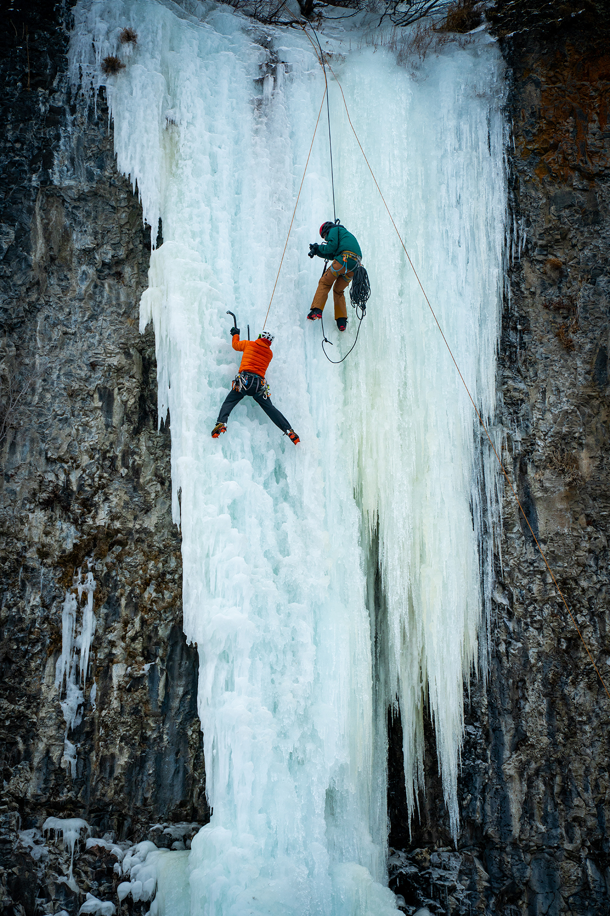 Winter sports photography of an ice climber ascending a frozen waterfall while a photographer captures the action from above on a rope.