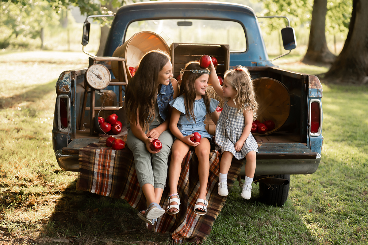 Três meninas compartilham maçãs e risadas na traseira de um caminhão antigo durante a golden hour - um cenário ideal para fotografar retratos de família com carinho. ©Marcie Reif