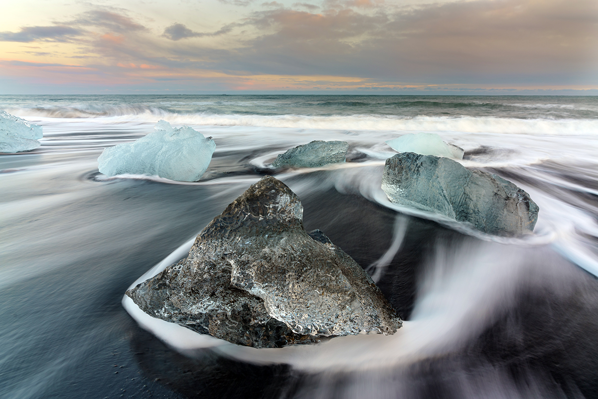 La orilla de una playa de arena negra en Islandia con grandes rocas en primer plano fotografiada utilizando fotografía de larga exposición para que las olas parezcan suaves.