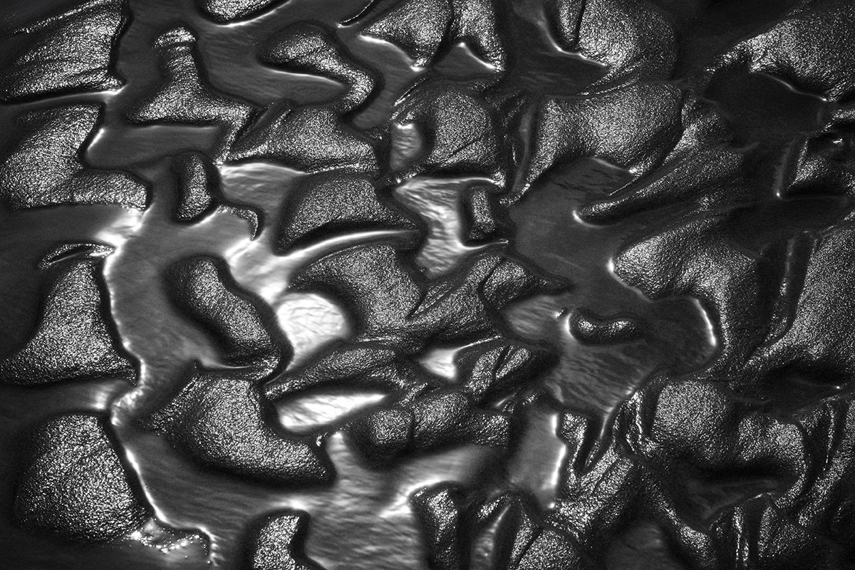 Close-up monocromático de padrões texturizados de areia úmida, demonstrando técnicas de fotografia abstrata por meio de luz, sombra e forma natural.