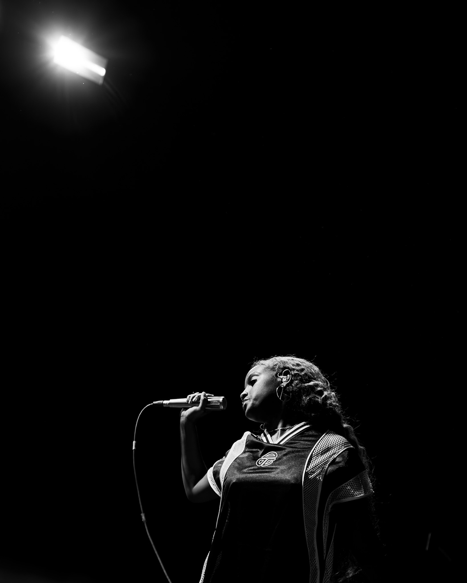 Fotografia de concerto em preto e branco de uma vocalista se apresentando sob um holofote, capturando a emoção e a intensidade da música ao vivo em um cenário minimalista.