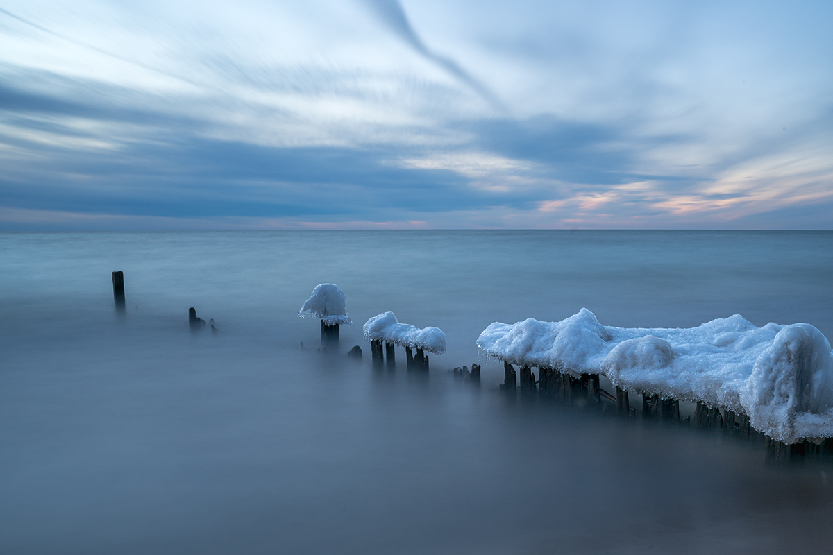 Exposição longa de postes de madeira gelados ao longo de uma costa de inverno ao pôr do sol, demonstrando dicas de fotografia de inverno para capturar paisagens congeladas.