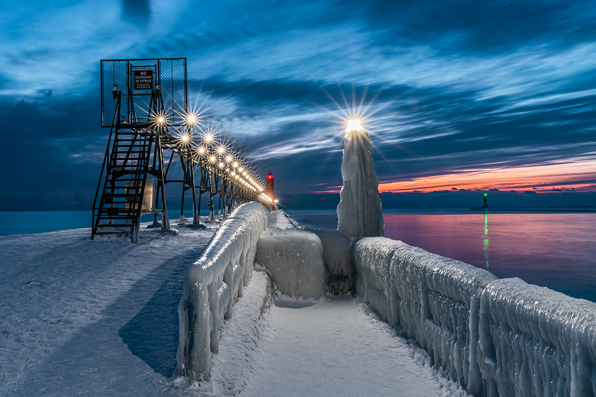 Píer congelado e farol iluminado ao pôr do sol, mostrando dicas de fotografia de inverno para capturar paisagens geladas e iluminação dramática.